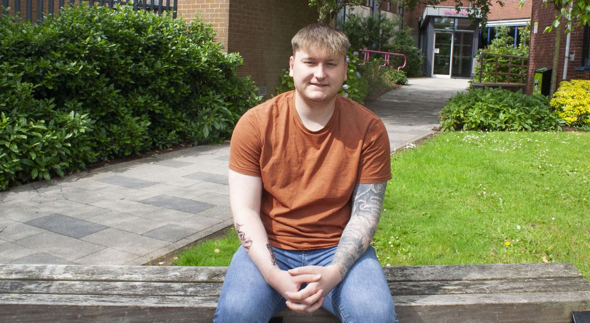 Chris Billington, young male wearing orange teeshirt and blue jeans sitting on a bench outside SERC's Bangor Campus smiling to camera.